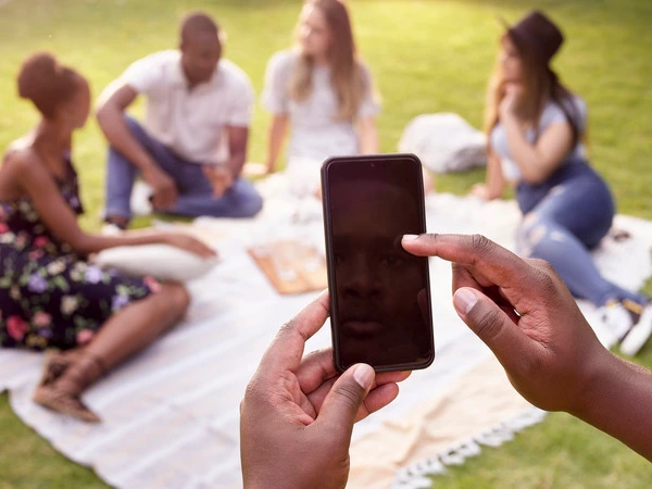 Group of people sitting on a picnic blanket with someone taking a photo with a mobile phone